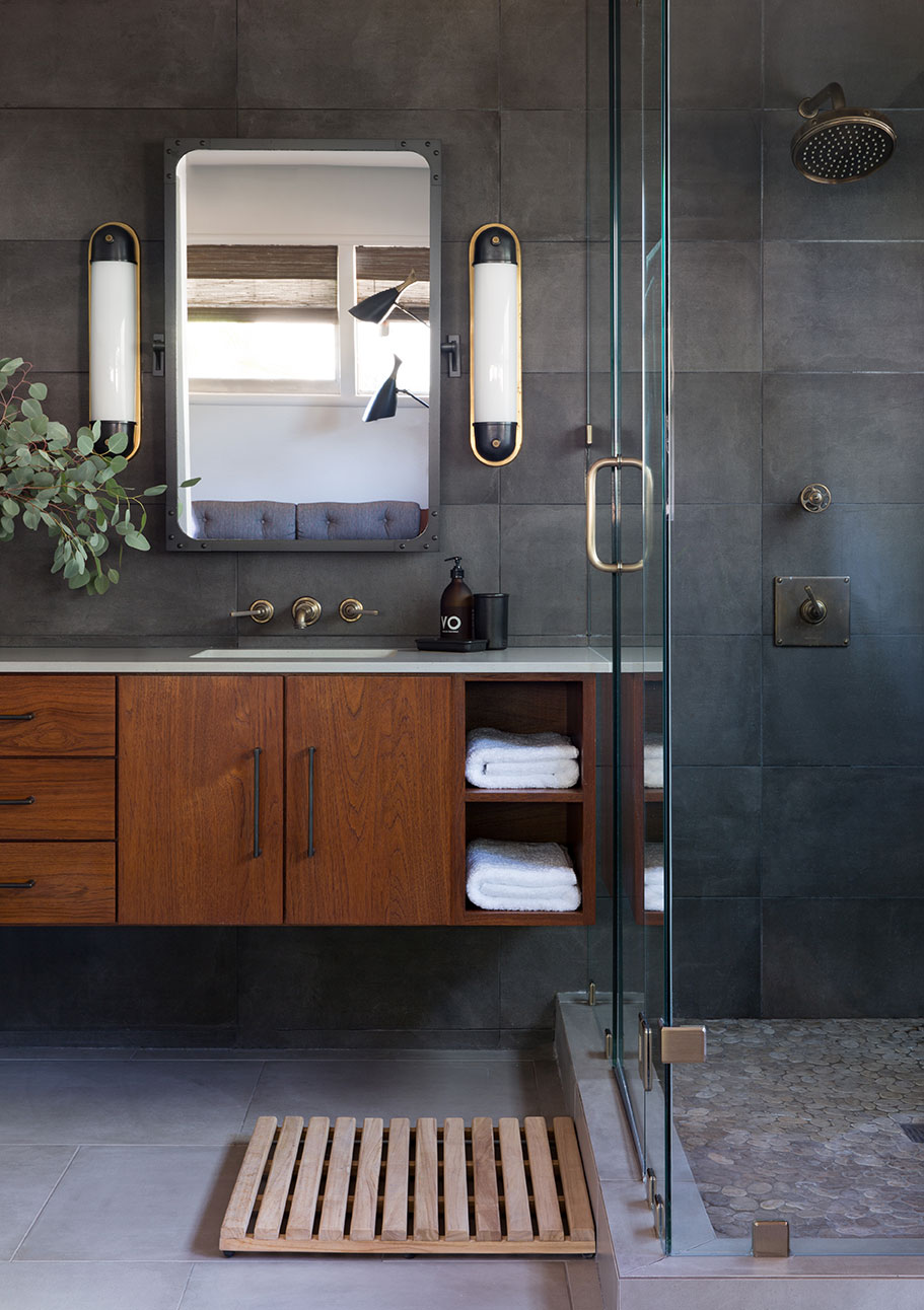 Leslie Phillips-Greco designed this grey bathroom with white counter tops, black and gold light fixtures, wood cabinets and a clear glass shower.