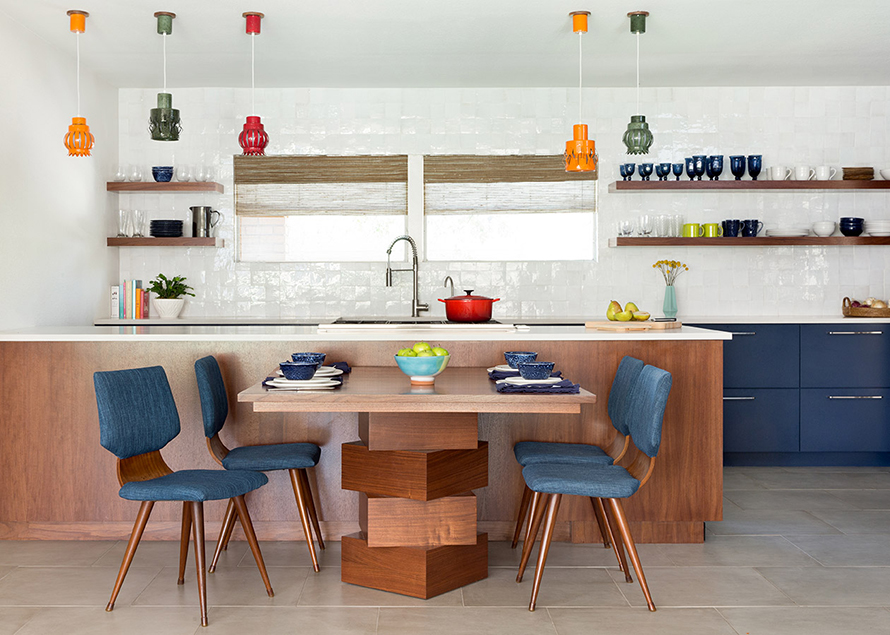 Beautiful open kitchen with blue cabinets, dark wood open shelving, colorful light fixtures, white counter tops and beautiful white tile backsplash with a unique wood table designed by Round Table Design.
