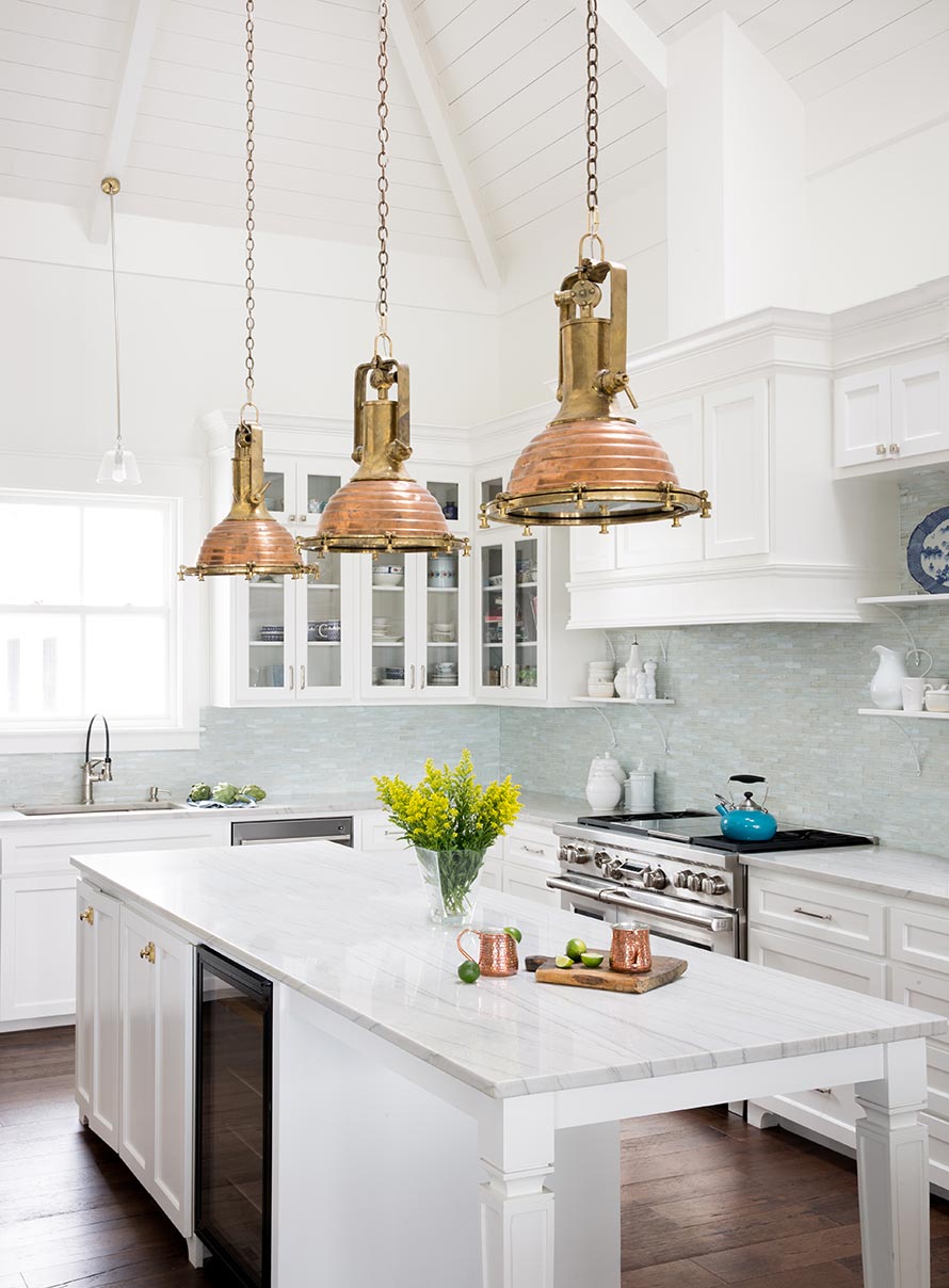 Open kitchen with white counter tops, white walls, white cabinets and light blue tile backsplash with antique gold hanging light fixtures.
